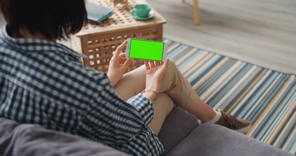 Young Woman Using Smartphone with Green Mock-up Screen Sitting on Sofa at Home