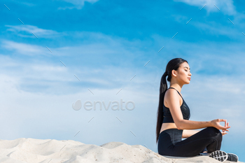 asian woman meditating in lotus yoga pose with gyan mudra on sand against cloudy sky / Photodune, Misc