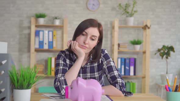 Young Woman Sitting at the Table Puzzled Looks at the Piggy Bank