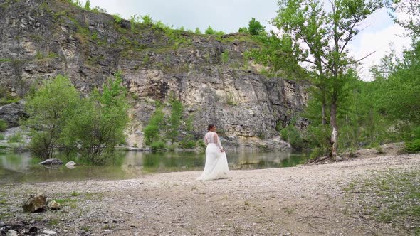 Bride in a White Dress with a Bouquet Walks on the Shore of a Mountain Lake