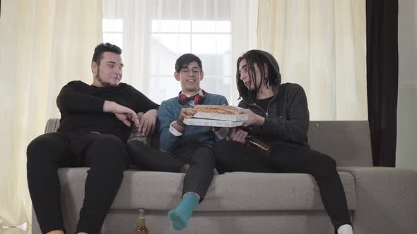 Three Caucasian Young Men Eating Pizza and Drinking Beer Indoors. Male University Students Sitting