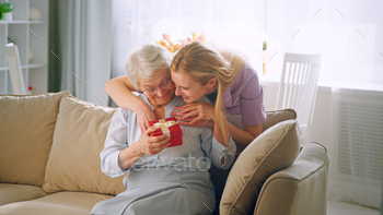 Long haired blonde in purple dress gives small box with present to aged lady / Photodune, Misc