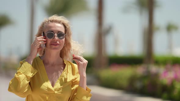 Portrait of a Blonde in Sunglasses and a Yellow Dress Against a Background of Blurred Palm Trees on