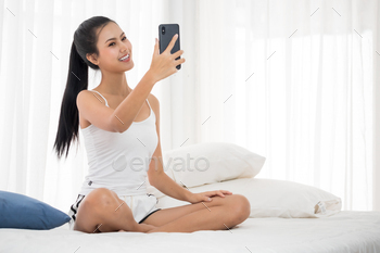 Beautiful asian woman smiling at a smartphone whlie sitting on the bed. / Photodune, Misc