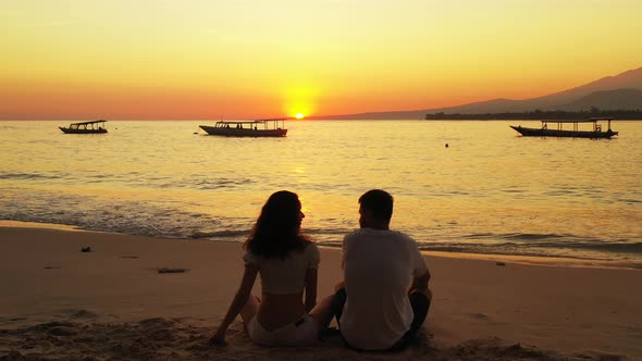 Couple sitting on exotic beach at beautiful sunset with yellow orange sky reflecting on calm sea sur
