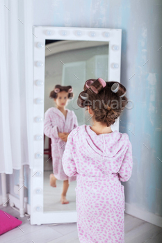 A child in bathrobe and curlers looking in mirror. The concept of fashion and beauty / Photodune, Misc