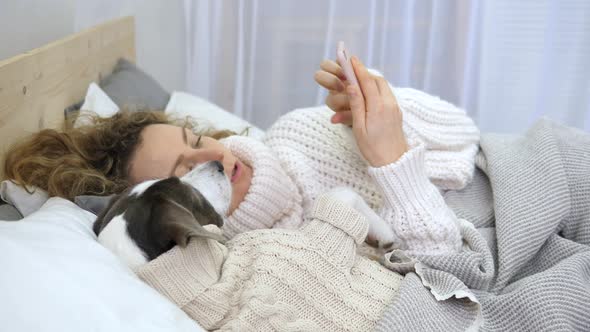 Young Woman Lying With Dog In Cozy Bed And Using Smartphone