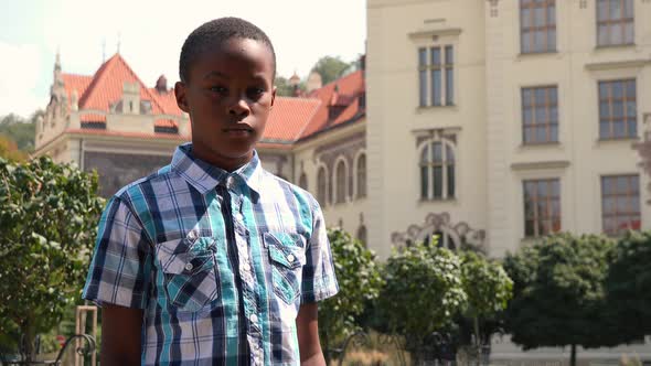 A Young Black Boy Looks Seriously at the Camera in an Urban Area - School in the Background