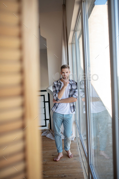 A man in jeans and a plaid shirt standing near the big window at home / Photodune, Misc
