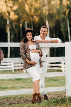 a pregnant girl in a hat and her husband in white clothes stand next to a horse corral at sunset.a / Photodune, Misc