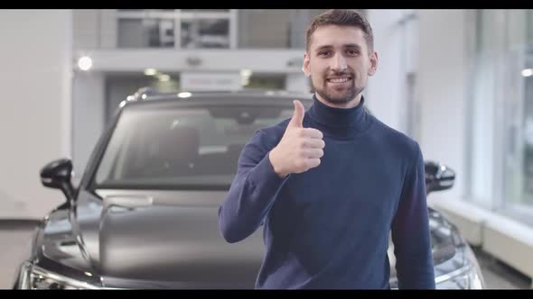 Portrait of Young Caucasian Man Standing in Front of New Car and Showing Thumb Up