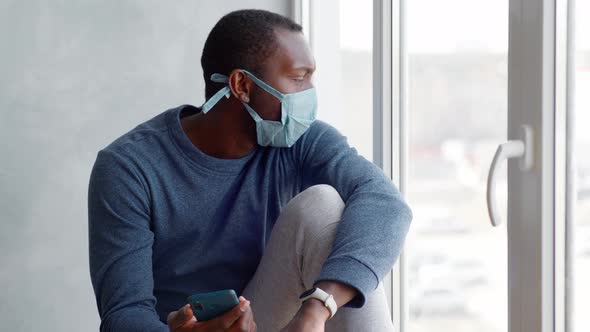 a Man with a Protective Mask on His Face During Quarantine Sits By the Window with a Smartphone