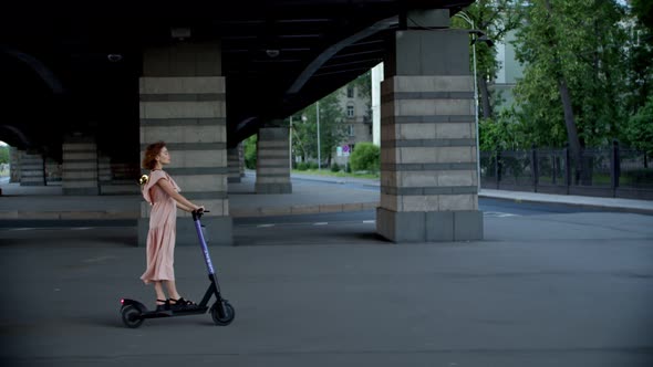 a Girl in a Dress Rides a Scooter Under the Bridge