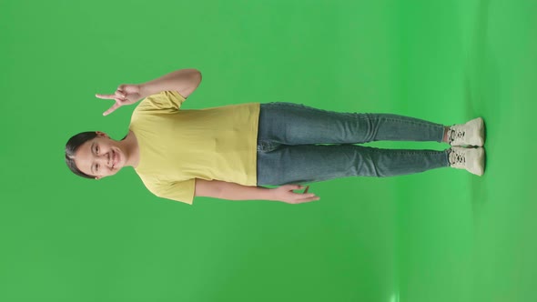 Full Body Of Smiling Young Asian Kid Girl Showing Gesture Peace While Standing On Green Screen