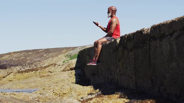 Senior african american man taking a break using smartphone on rocks by the sea