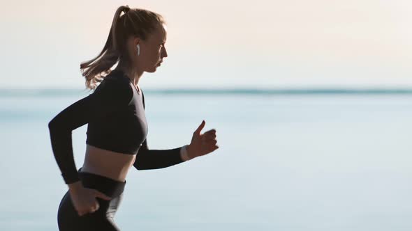 Active Young Sportswoman in Earphones Running on Beach at Sunset