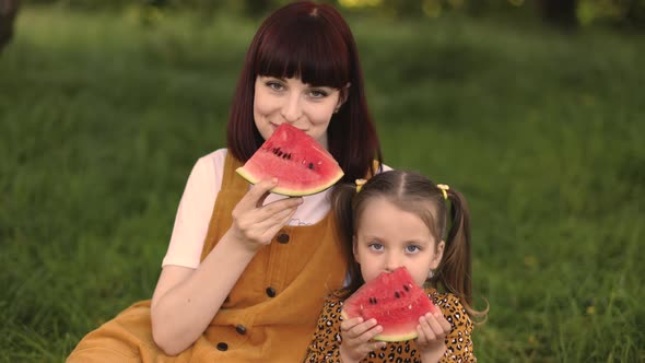 Young Mother Resting with Her Daughter in Park Touch the Noses and Holding a Watermelon