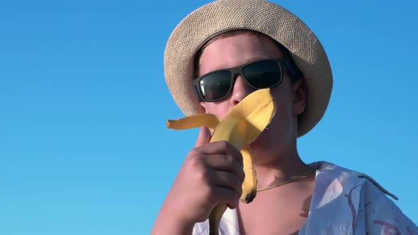Boy in a Hat and Sunglasses Eats a Banana Against Blue Sky Background