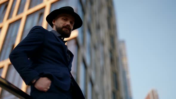 Confident Elegant Man in Suit and Hat is Standing Against Modern High Building Tilt Up View