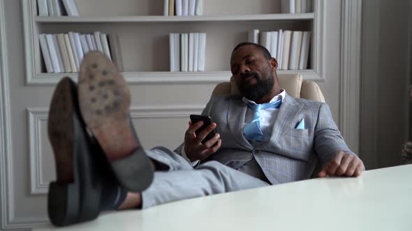 Rich Black Man Is Dressed Expensive Business Suit Is Sitting in Armchair and Holding Legs on Table