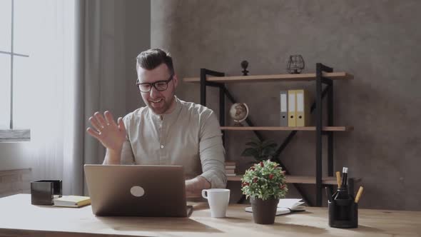 Positive Man in Glasses Dressed Casual , Having Video Call Over Laptop, Waving and Smiling. On Table