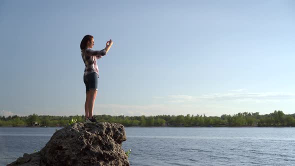 A Young Woman Stands on a Stone Near the River and Photographs the Landscape. The Girl Takes