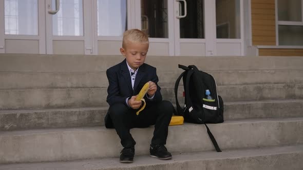 Schoolboy Sits on the Stairs at School and Eats a Banana, Next To Is a Backpack