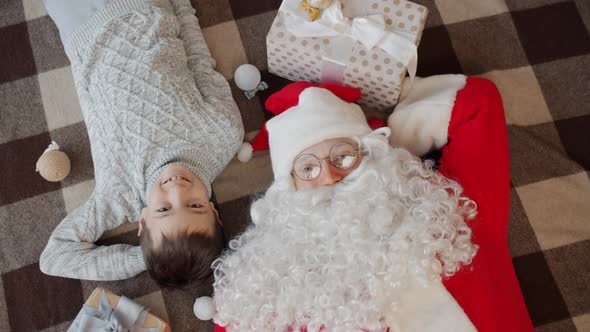 Portrait of Kid and Santa Claus Looking at Camera Lying on Floor at Home