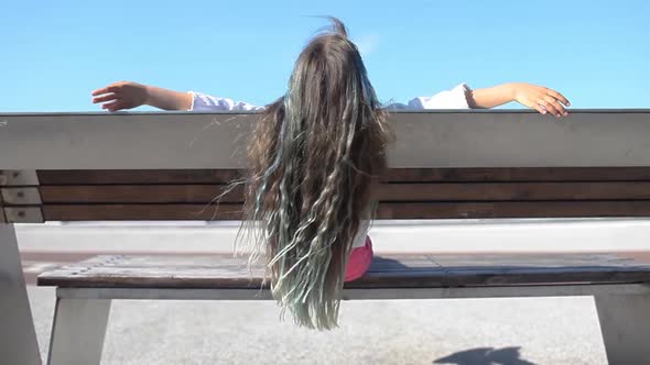 Brunette Preteen Girl with Blue Color Highlights Sitting on Bench on Bridge Back View Summer Day