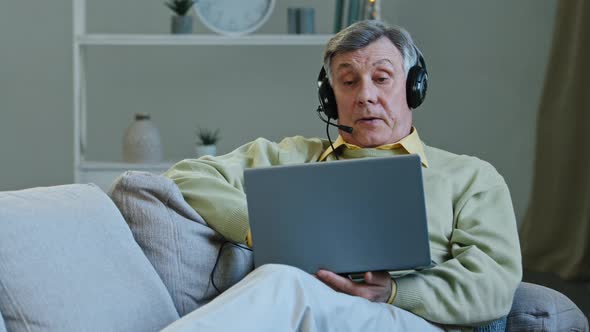 Senior 60s Male on Couch at Home Wear Digital Earphones Talking Online to Laptop Web Camera Old Man