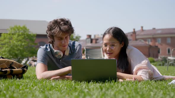 Cheerful Young University Students Lying on Green Grass in Park and Using Laptop