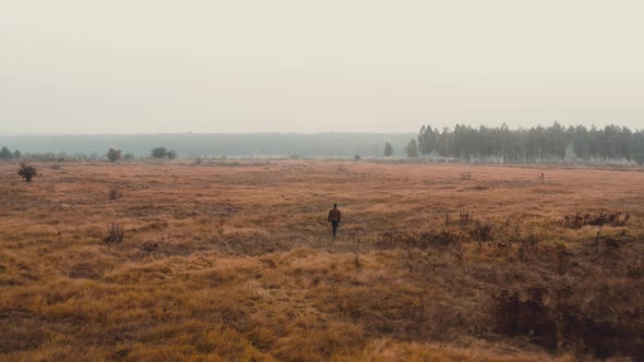 Man carrying a camera walking in a misty autumn field towards a forest.
