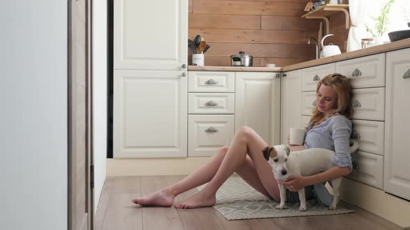 Pregnant Woman in Pajamas Sitting on the Floor in the Kitchen. Woman in Shorts and a Shirt Drinks