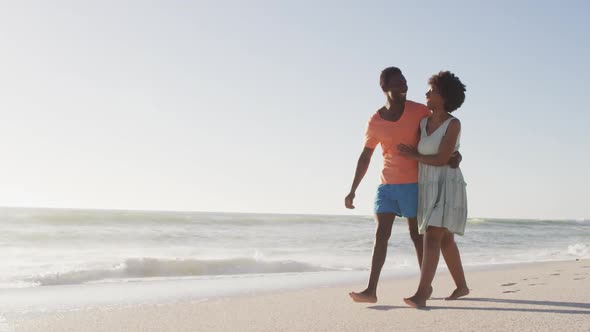 Smiling african american couple embracing and walking on sunny beach