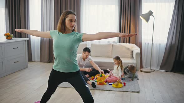 Confident Mother Practicing Yoga Standing in Warrior Pose While Her Family Playing on Background