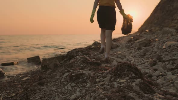 A female volunteer walks along the beach with a full bag of garbage and collects plastic bottles