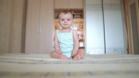 Adorable Baby with Blue Eyes Sits on the Bed. A Toddler Smiles and Laughs at the End. White Clothes