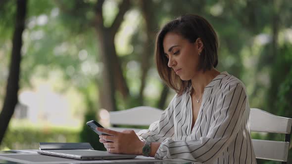 Young Hispanic Latin Ethnic Woman Sitting in a Summer Cafe Holding Phone Looking at Screen Watching