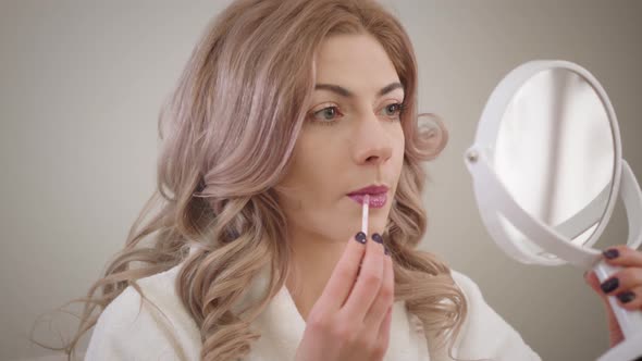 Close-up Portrait of Charming Caucasian Girl Looking at Small Round Mirror and Applying Lipstick