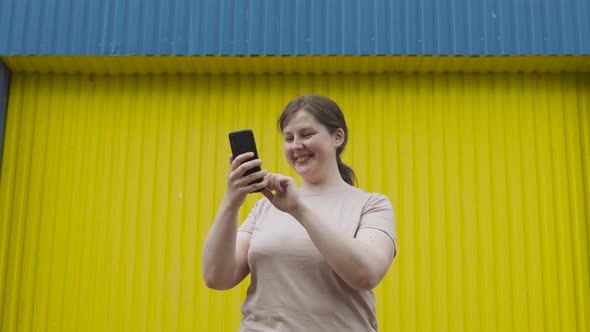 Positive Simple Young Woman Smiling and Using Smartphone Near Yellow Blue Wall