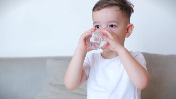 Cute Baby Boy Drinking a Glass of Water Sitting on the Couch at Home. Slow Motion Little Boy
