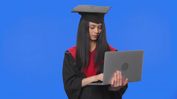 Portrait of Female Student in Cap and Gown Graduation Costume Communicates Online Using a Laptop