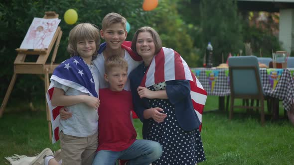 Positive Boys and Girl Wrapped in American Flag Posing on Spring Summer Backyard Smiling