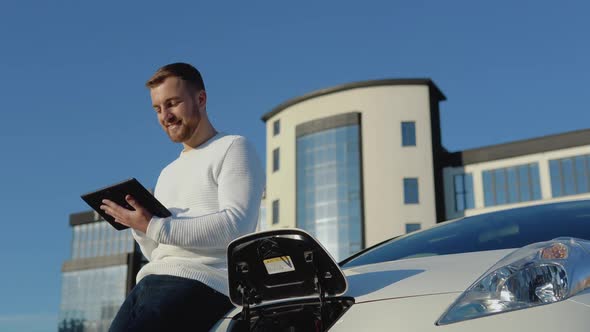 A Male Driver Stands with a Tablet in His Hands Near an Electric Car While It is Charging