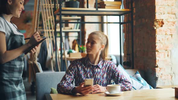 Pretty Girl Using Smartphone in Coffee House Ordering From Friendly Waitress