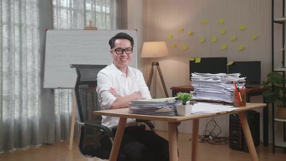 Asian Man Crossing Arms And Smiling To Camera While Working With Documents At The Office