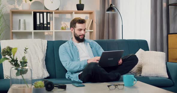 Bearded Man Sitting in Lotus Pose on the Couch at Home and Working on Computer