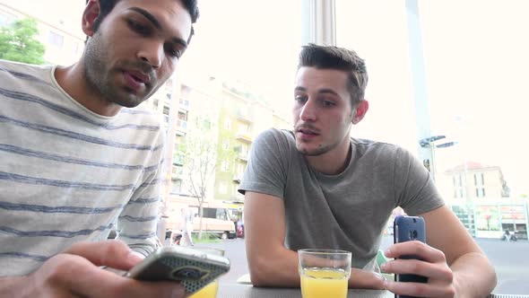 two young multi ethnic men outdoor sitting bar using smart phone having conversation