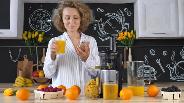 Female Drinking Orange Juice In Kitchen And Using Smartphone