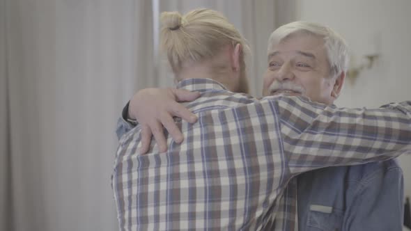 Close-up of Happy Elderly Caucasian Man Hugging Adult Son Indoors. Portrait of Smiling Senior